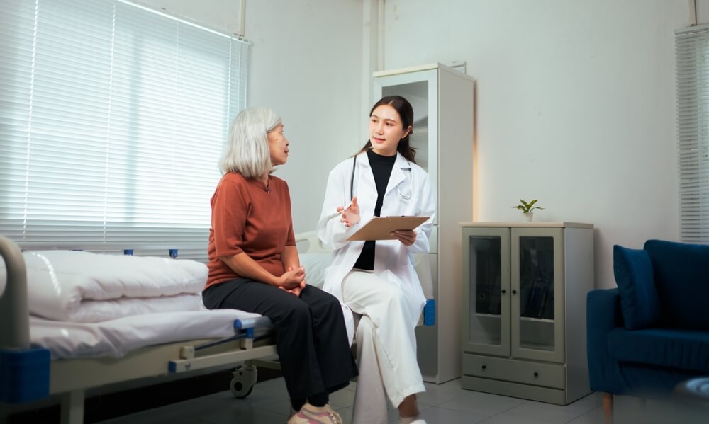 Asian female doctor discussing medical information with a senior woman sitting on an examination bed.