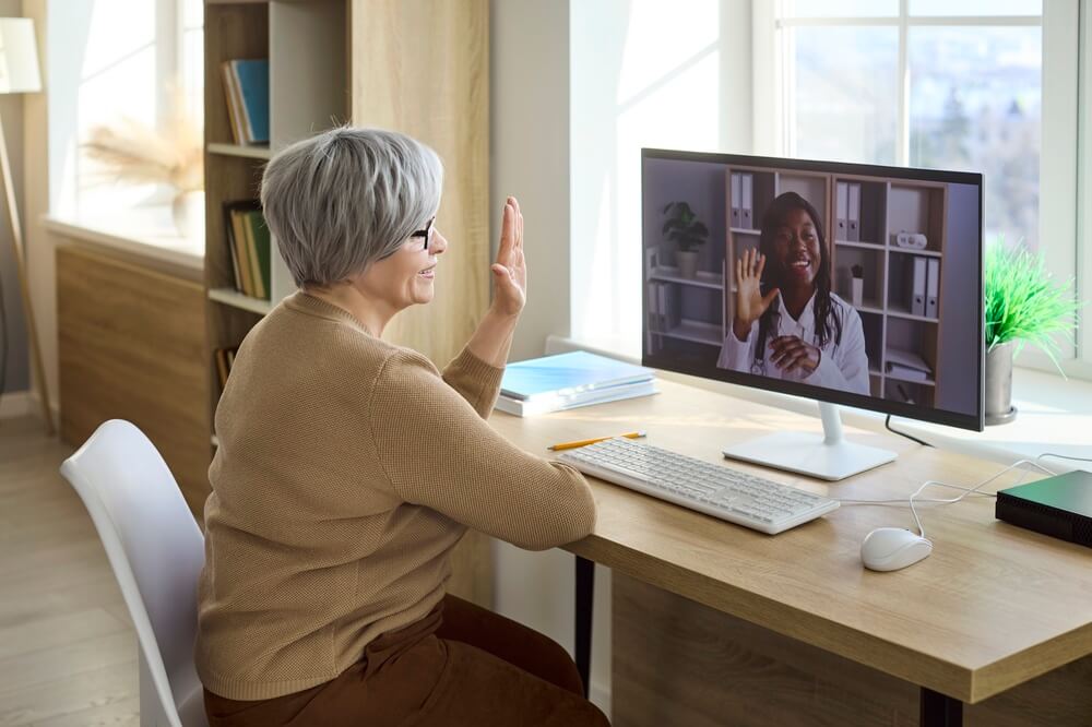 Doctor and senior woman telemedicine video consultation online. An elderly patient greets a doctor on a computer during a remote care call from home. Modern telehealth for seniors at home.